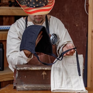 May include: A person holding a black quilted hood and a cream-colored quilted hood with black ties. The person is wearing a straw cowboy hat with an American flag design and a brown apron.