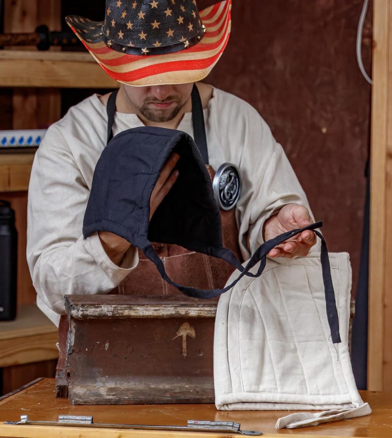 May include: A person holding a black quilted hood and a cream-colored quilted hood with black ties. The person is wearing a straw cowboy hat with an American flag design and a brown apron.