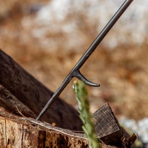 May include: A close-up of a dark metal fire poker with a pointed end and a curved hook. The poker is angled downward, resting on a weathered wooden surface, with a blurred natural background.