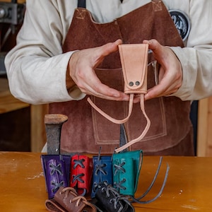 May include: A light brown leather sheath held by a person, with two small black rivets and long leather ties. Below are several colourful leather sheaths in purple, red, blue, teal, brown, and black, each with decorative lacing.
