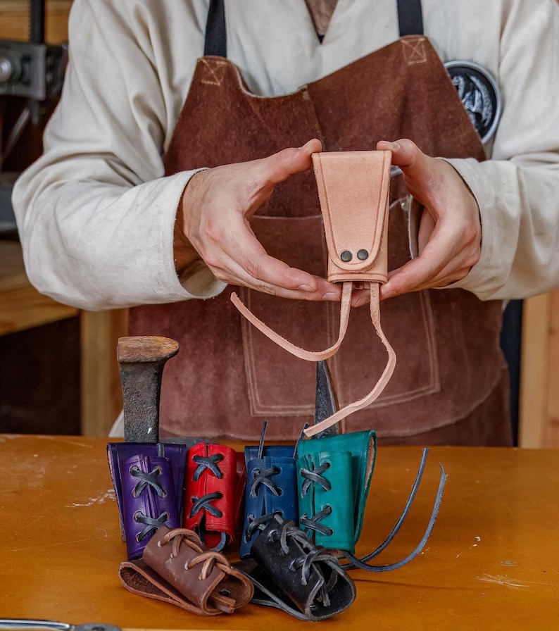 May include: A light brown leather sheath held by a person, with two small black rivets and long leather ties. Below are several colourful leather sheaths in purple, red, blue, teal, brown, and black, each with decorative lacing.