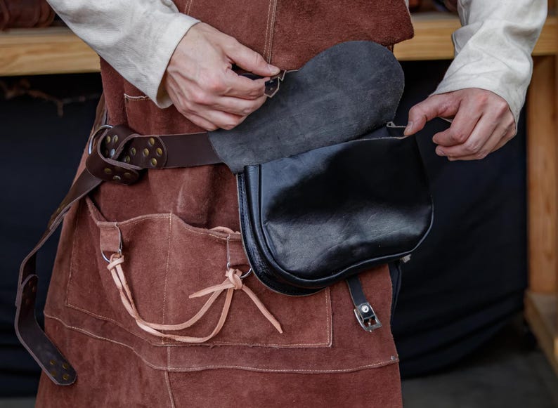 Op de afbeelding: Een bruine leren riem met een gesp en een zwarte leren tas eraan bevestigd. De tas heeft een klep die naar beneden is geklapt.