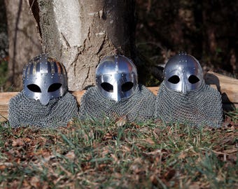 May include: Three polished metal helmets with eye openings and chainmail neck protection are displayed outdoors. The helmets have a medieval design, with rivets and curved visors. They are set against a backdrop of a tree trunk and foliage.
