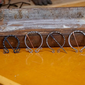 May include: Four handcrafted metal brooches with unique designs. The brooches are silver-toned with decorative elements and copper-colored accents. They are displayed on a yellow surface with a weathered metal tray in the background. Each brooch is approximately 2 inches in diameter.