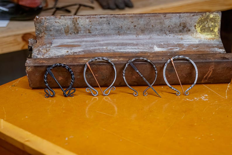 May include: Four handcrafted metal brooches with unique designs. The brooches are silver-toned with decorative elements and copper-colored accents. They are displayed on a yellow surface with a weathered metal tray in the background. Each brooch is approximately 2 inches in diameter.
