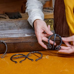 May include: Hand-forged, dark metal brooches in various designs. One brooch is held by a person, showcasing its intricate spiral and looped details. The brooches are displayed on a yellow surface, suggesting a craft or historical theme.