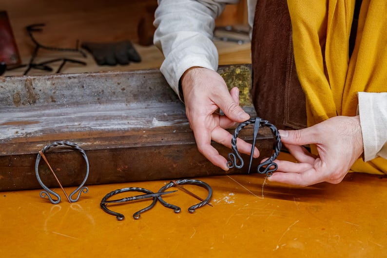 May include: Hand-forged, dark metal brooches in various designs. One brooch is held by a person, showcasing its intricate spiral and looped details. The brooches are displayed on a yellow surface, suggesting a craft or historical theme.