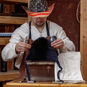 May include: A person wearing a cowboy hat with a red, white, and blue American flag design. The person is working on a black and navy blue padded hood, with a white quilted hood nearby. The person is wearing a white shirt and brown apron.