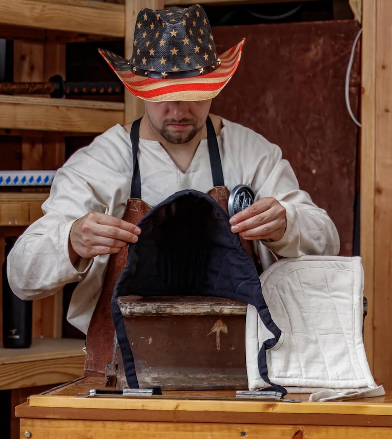 May include: A person wearing a cowboy hat with a red, white, and blue American flag design. The person is working on a black and navy blue padded hood, with a white quilted hood nearby. The person is wearing a white shirt and brown apron.