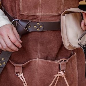 Puede incluir: Un delantal de cuero marr&oacute;n con un bolsillo grande y una bolsa m&aacute;s peque&ntilde;a unida al cintur&oacute;n. El cintur&oacute;n tiene remaches de lat&oacute;n y una hebilla de metal. El delantal lo lleva puesto una persona, pero se ven las manos de la persona.