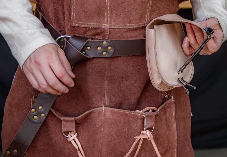 Puede incluir: Un delantal de cuero marr&oacute;n con un bolsillo grande y una bolsa m&aacute;s peque&ntilde;a unida al cintur&oacute;n. El cintur&oacute;n tiene remaches de lat&oacute;n y una hebilla de metal. El delantal lo lleva puesto una persona, pero se ven las manos de la persona.