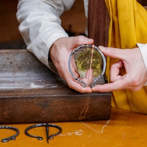 May include: A handcrafted, circular brooch with a copper pin, held by hands. The brooch is made of dark metal with decorative swirls. Other similar brooches are visible on a wooden surface.