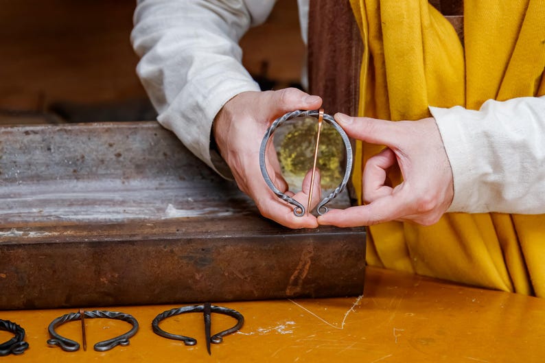 May include: A handcrafted, circular brooch with a copper pin, held by hands. The brooch is made of dark metal with decorative swirls. Other similar brooches are visible on a wooden surface.
