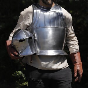 Pu&ograve; includere: Una corazza in acciaio lucido con nervature verticali sul petto superiore e un elmo in acciaio abbinato con un'apertura di ventilazione a forma di croce. L'armatura &egrave; indossata sopra una camicia a maniche lunghe con guanti in pelle marrone.