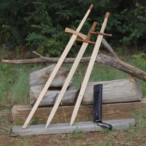 May include: Three wooden swords with light-colored blades and dark brown crossguards and pommels. The swords feature straight, tapered blades and are accompanied by a black rectangular carrying case with an adjustable strap.