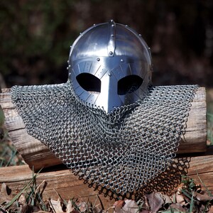 May include: A polished steel Viking helmet with eye and nose openings, riveted construction, and a chainmail coif. The helmet and chainmail are displayed on a weathered wooden log, with a blurred natural background.