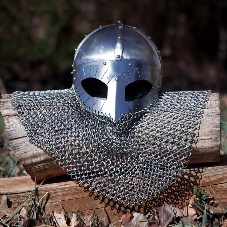 May include: A polished steel Viking helmet with eye and nose openings, riveted construction, and a chainmail coif. The helmet and chainmail are displayed on a weathered wooden log, with a blurred natural background.