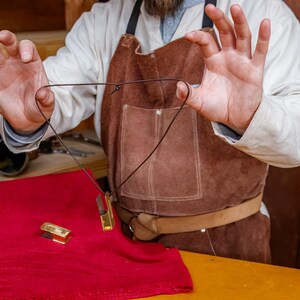 Pu&ograve; includere: Una collana con pendente dorato e cordino nero &egrave; mostrata su un tessuto rosso. Un oggetto rettangolare dorato abbinato &egrave; sul tessuto. La persona indossa un grembiule di pelle marrone e una camicia bianca.