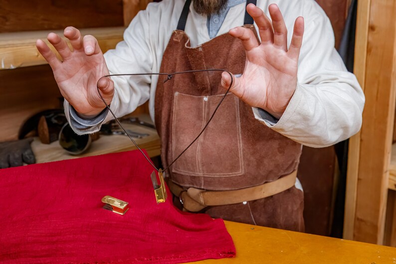 Pu&ograve; includere: Una collana con pendente dorato e cordino nero &egrave; mostrata su un tessuto rosso. Un oggetto rettangolare dorato abbinato &egrave; sul tessuto. La persona indossa un grembiule di pelle marrone e una camicia bianca.
