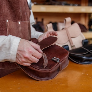 Op de afbeelding: Een persoon houdt een bruine leren tas vast met een gespsluiting. De tas wordt vastgehouden voor een houten plank met andere leren tassen erop.