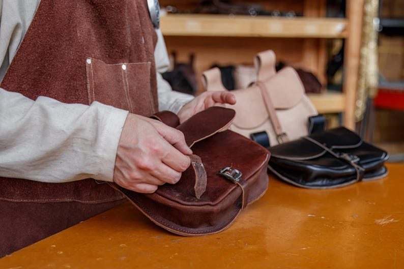 Op de afbeelding: Een persoon houdt een bruine leren tas vast met een gespsluiting. De tas wordt vastgehouden voor een houten plank met andere leren tassen erop.