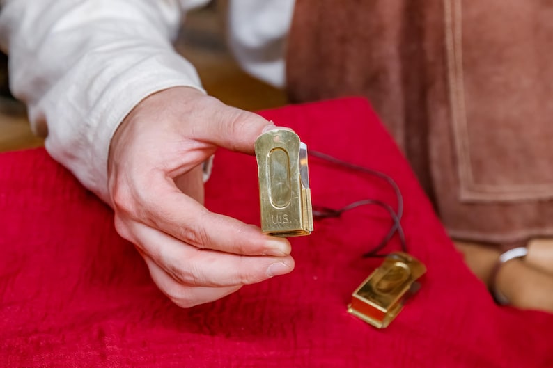 Pu&ograve; includere: Un paio di fermasoldi in metallo color oro, uno tenuto in mano, l'altro su una superficie rossa. La clip in mano ha incise le lettere "US". Un cordino scuro &egrave; attaccato a ciascuna clip.