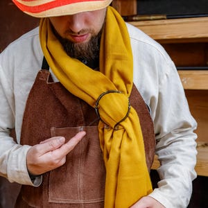 May include: A mustard yellow scarf is looped through a dark metal ring. The scarf is draped over a white shirt and brown apron. A straw hat with red and white stripes is worn.