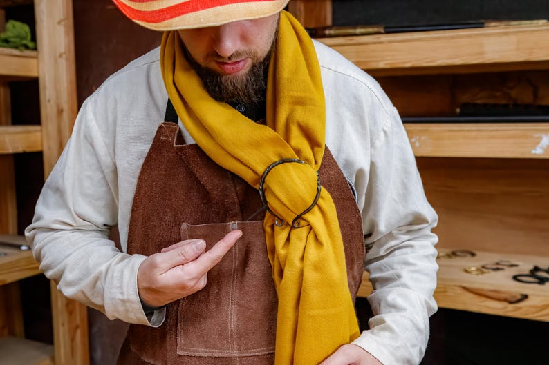 May include: A mustard yellow scarf is looped through a dark metal ring. The scarf is draped over a white shirt and brown apron. A straw hat with red and white stripes is worn.