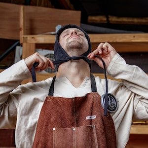 May include: A person wearing a black hood and a brown leather apron with a white shirt underneath. The person is tying the hood's strings. The apron has a pocket and a metal badge. The background is a wooden shelf.