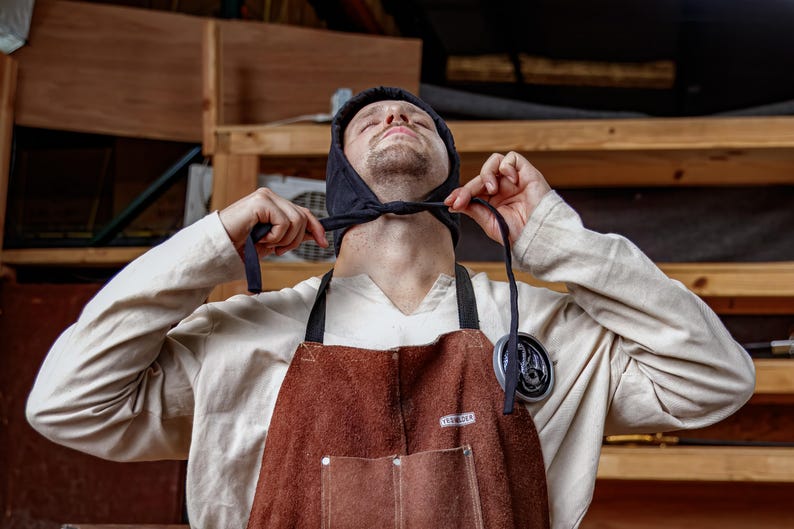 May include: A person wearing a black hood and a brown leather apron with a white shirt underneath. The person is tying the hood's strings. The apron has a pocket and a metal badge. The background is a wooden shelf.