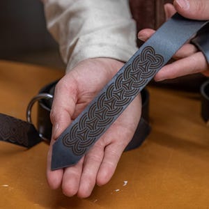 May include: A close-up of a dark blue leather belt with a detailed Celtic knot design. The belt is held in a person's hand, showing the intricate details of the pattern.