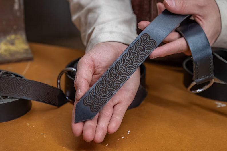 May include: A close-up of a dark blue leather belt with a detailed Celtic knot design. The belt is held in a person's hand, showing the intricate details of the pattern.