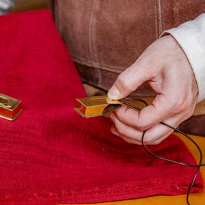 Pu&ograve; includere: Due piccoli oggetti rettangolari in metallo dorato con interno rosso sono su un panno rosso. Una mano tiene uno degli oggetti, infilando un cordoncino nero. Gli oggetti sembrano decorativi o funzionali.