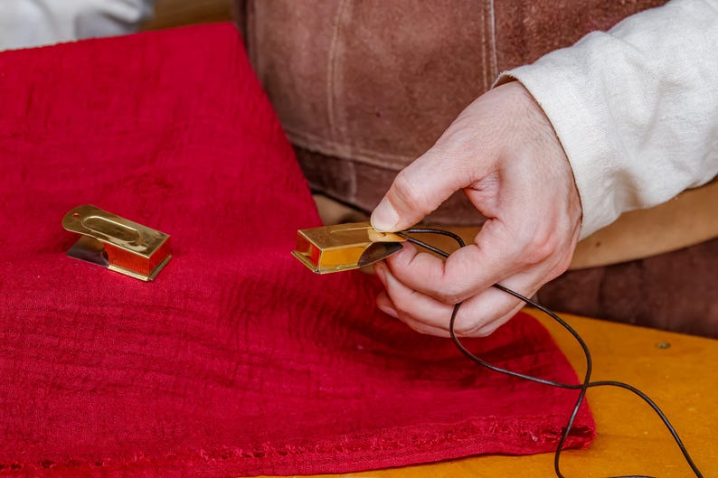 Pu&ograve; includere: Due piccoli oggetti rettangolari in metallo dorato con interno rosso sono su un panno rosso. Una mano tiene uno degli oggetti, infilando un cordoncino nero. Gli oggetti sembrano decorativi o funzionali.