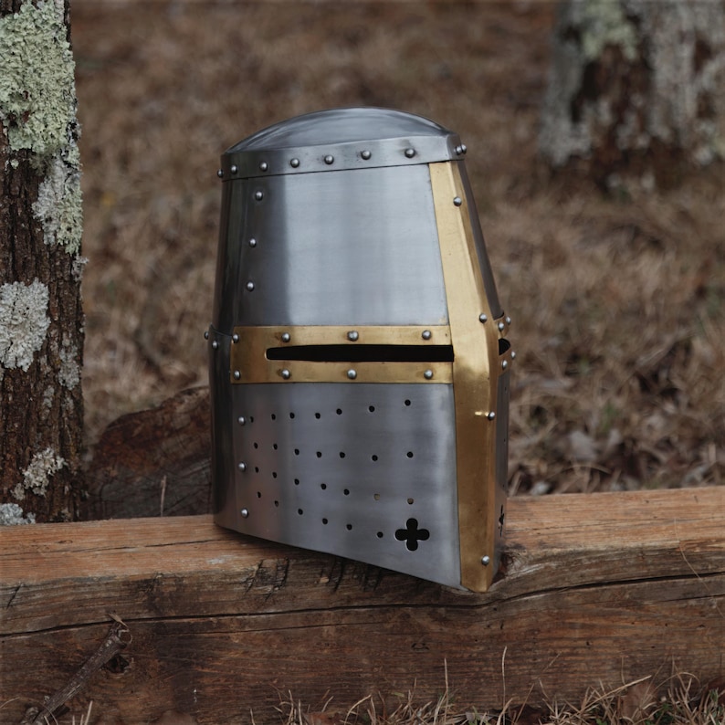 May include: A silver riveted helmet with brass detailing. The helmet has a cross design and is likely historical or medieval in style. It is displayed on a wooden surface, suggesting a display or decorative item.