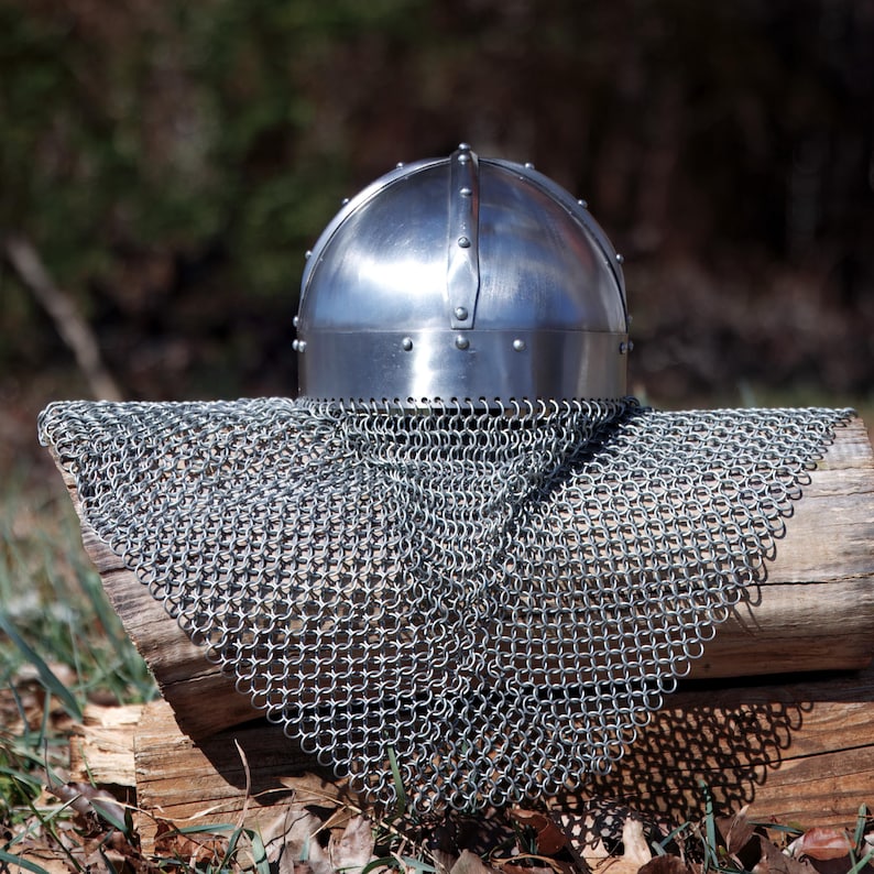 May include: A shiny steel helmet with rivets and a chainmail coif. The helmet rests on a weathered wooden log, with the chainmail draped over it. The background is blurred, suggesting an outdoor setting.