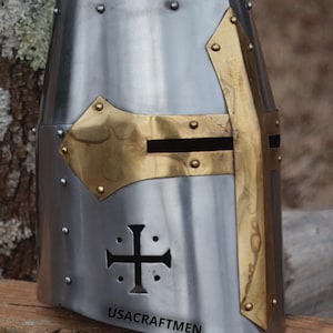 May include: A silver medieval helmet with brass detailing. The helmet has a cross design on the front and brass accents around the face opening. The text "USACRAFTMEN" is printed on the lower front.