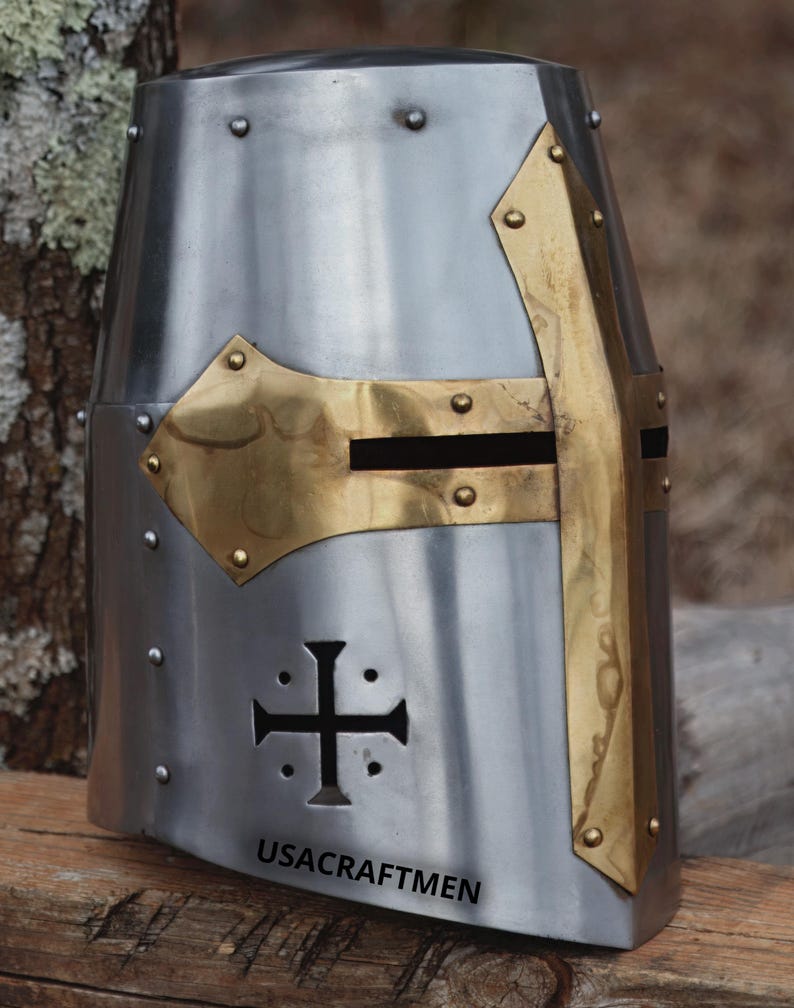 May include: A silver medieval helmet with brass detailing. The helmet has a cross design on the front and brass accents around the face opening. The text "USACRAFTMEN" is printed on the lower front.