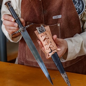 May include: A silver and black dagger with a leather sheath. The dagger has a long, sharp blade and an ornate handle. The sheath is tan leather with a decorative metal end. The dagger is held by a person wearing a brown apron.