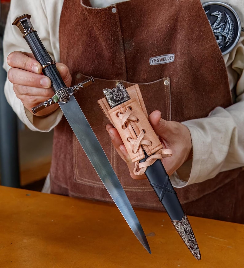 May include: A silver and black dagger with a leather sheath. The dagger has a long, sharp blade and an ornate handle. The sheath is tan leather with a decorative metal end. The dagger is held by a person wearing a brown apron.