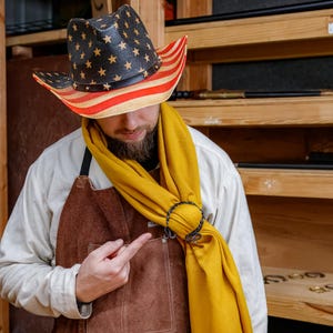 May include: A straw cowboy hat with a black crown and a red and white striped brim, adorned with gold stars. The hat is worn with a yellow scarf and a brown leather apron.