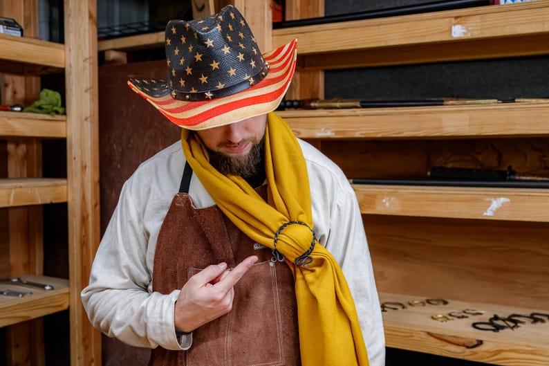 May include: A straw cowboy hat with a black crown and a red and white striped brim, adorned with gold stars. The hat is worn with a yellow scarf and a brown leather apron.