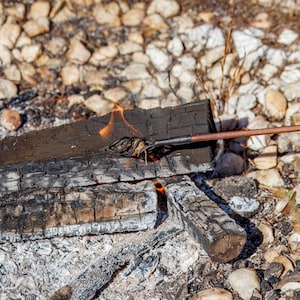 May include: A close-up shot of a campfire with burning wood and flames. A metal branding iron is being used to brand something on the wood. The wood is charred black and surrounded by rocks.