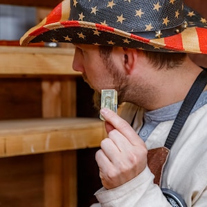 Pu&ograve; includere: Un cappello da cowboy di paglia con un design patriottico con strisce rosse e bianche e blu con stelle bianche. La persona che indossa il cappello tiene in mano una piccola pila di banconote da un dollaro USA.