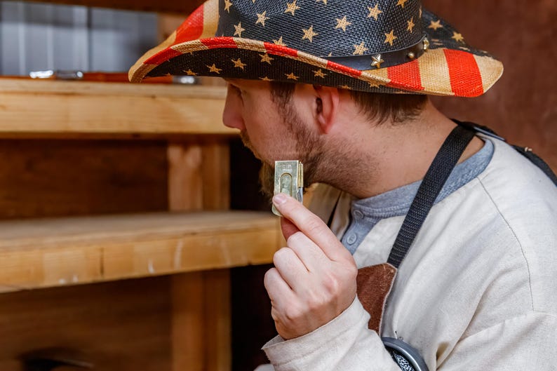 Pu&ograve; includere: Un cappello da cowboy di paglia con un design patriottico con strisce rosse e bianche e blu con stelle bianche. La persona che indossa il cappello tiene in mano una piccola pila di banconote da un dollaro USA.