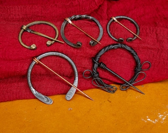 May include: A collection of five metal brooches with varying designs. The brooches are circular or semi-circular, with a pin running across. The metal colors include bronze, silver, and black, set against a red fabric background.