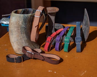 May include: A set of seven leather straps with silver buckles in various colors, including red, purple, green, and brown. The straps are arranged on a wooden surface next to a horn drinking mug.