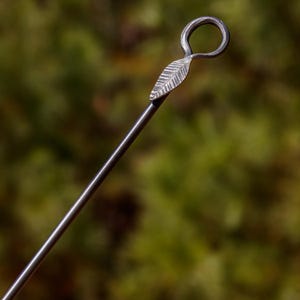 May include: A close-up of a metal skewer with a leaf design. The skewer has a loop at the top and a leaf-shaped detail. The skewer is made of a dark metal and is set against a blurred green background.