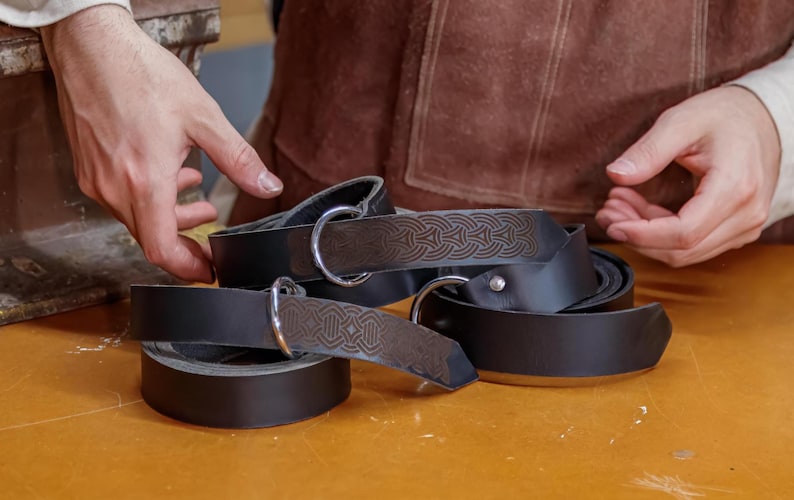 May include: A set of three black leather belts with silver buckles and intricate designs. The belts are stacked on top of each other and are lying on a wooden surface.