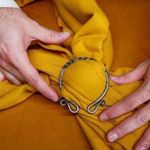 May include: A silver-colored metal cloak clasp with a woven design, set against a mustard yellow fabric. The clasp has a circular shape with decorative swirls. The fabric is being held by hands.